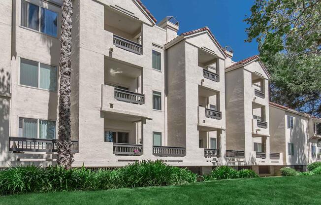 Three-story residential building with light-colored stucco walls, featuring multiple balconies and large windows. The surrounding area has lush green grass and landscaping, including palm trees. The clear blue sky creates a bright, inviting atmosphere.