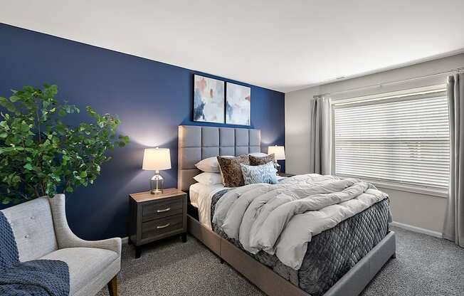 A bedroom with a bed, nightstand, chair, and a plant.