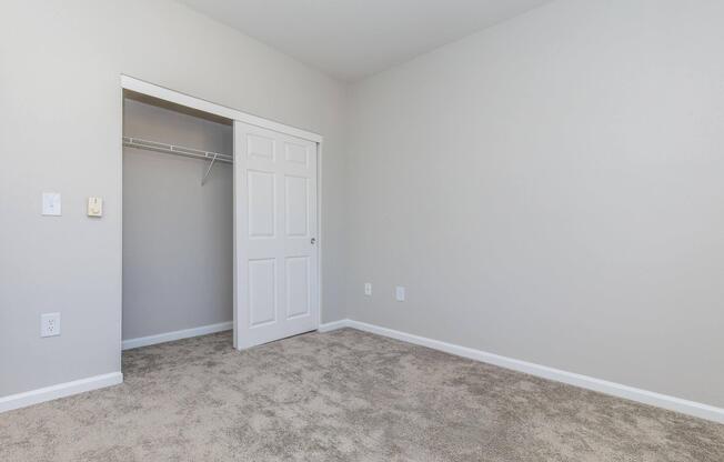 Empty room with light grey walls and a carpeted floor. A white door leads to a closet on the left, and the room has a minimalistic design with no furniture or decorations. The overall ambiance is bright and clean, suitable for various uses.