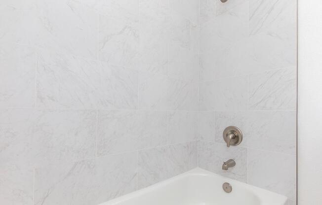 A modern bathroom featuring a white bathtub with a sleek chrome faucet and showerhead against a light marble tile backsplash. The walls are tiled with a light, textured marble pattern, contributing to a clean and contemporary aesthetic.