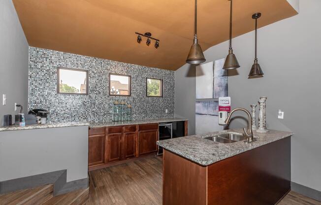 Modern kitchen interior featuring a granite countertop and wooden cabinets. The backsplash is adorned with blue mosaic tiles, and there are three small windows on the wall. Overhead, stylish pendant lights illuminate the space, while a sink and coffee maker are visible on the countertop.