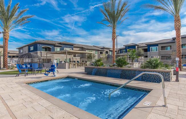 A modern outdoor pool area featuring a hot tub with a railing, surrounded by palm trees and lounge chairs. In the background, there are apartment buildings with balconies and a shaded seating area. The sky is bright blue with wispy clouds, creating a relaxing atmosphere.