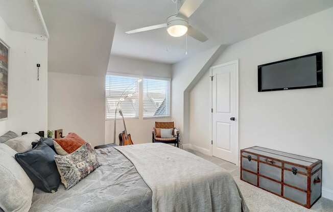 A bedroom with a bed, a guitar, a window with blinds, a ceiling fan, a wall-mounted TV, and a wooden nightstand.