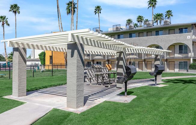 A shaded outdoor grilling area featuring two BBQ grills and a picnic table, surrounded by lush green grass and tall palm trees. Nearby is a multi-story building and a fenced area, creating a pleasant recreational space.