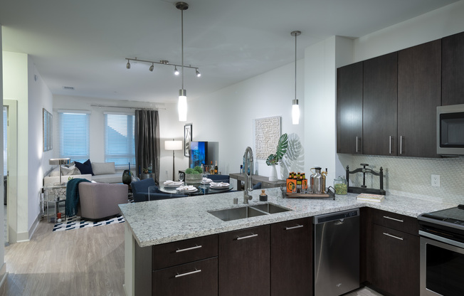 Open-concept kitchen, dining, and living space with ash-gray wood-style floors, dark modern wood cabinets, stainless steel appliances, granite countertops, and a spacious living area with windows and a glass door leading to the balcony.