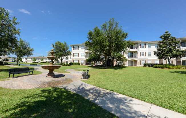 A park with a bench, a fountain and apartment buildings in the background.