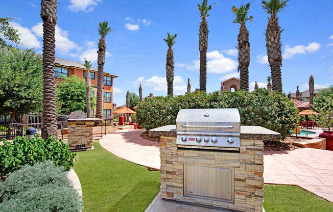 A landscaped outdoor grilling station here at Tuscany at Faudree featuring a built-in stainless steel grill on a stone base, surrounded by manicured green lawn, palm trees, and curved walkways. Mediterranean-inspired apartment buildings, poolside seating, and bright blue skies create a welcoming community atmosphere.