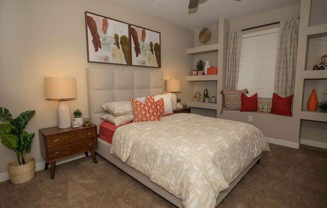 A cozy bedroom featuring a light-colored bed with decorative pillows, a stylish headboard, and wooden nightstands. There are two modern lamps on the nightstands, plants in a decorative pot, and wall art above the bed. The room is accented with orange and neutral tones, and has shelves displaying decorative items.