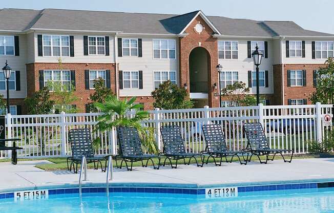 A pool in front of a brick building with a white fence and chairs.