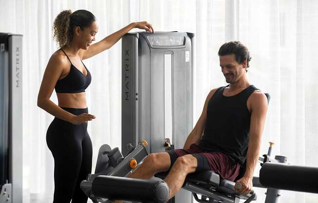 A woman is adjusting the settings on a gym machine while a man sits on a rowing machine. at Palma, Doral, FL