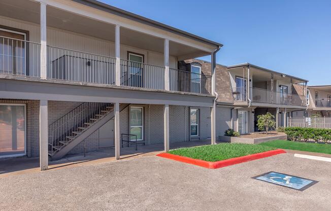 A multi-story apartment building exterior featuring a concrete parking lot. There are two levels of balconies, stairs leading to the upper level, and a designated wheelchair-accessible parking space in the foreground. The scene is under a clear blue sky, showcasing a well-maintained property.