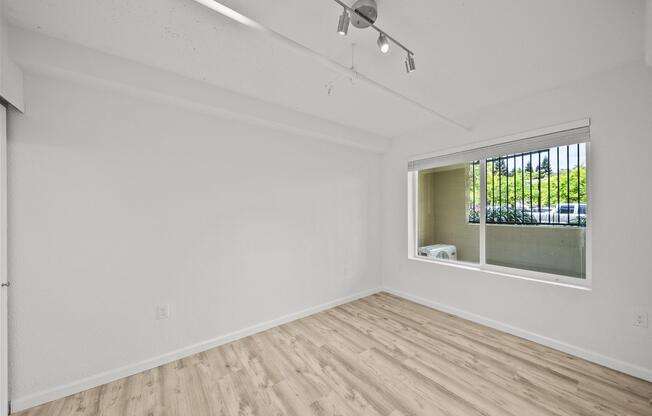 Interior of a modern, empty room featuring light-colored walls and wooden flooring. A large window allows natural light in, offering a view of green trees and an air conditioning unit outside. The ceiling is equipped with track lighting, enhancing the brightness of the space.