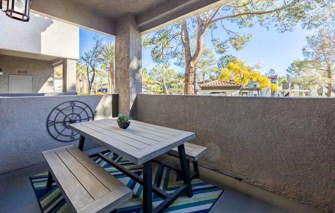 A table and bench are on a patio with a wall and trees in the background.