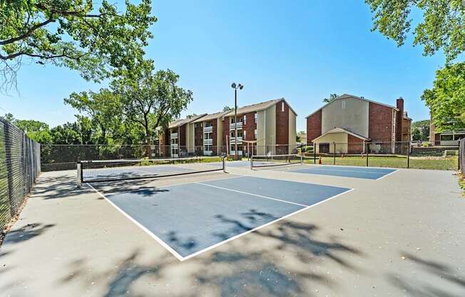 A tennis court is surrounded by a fence and there are buildings in the background.