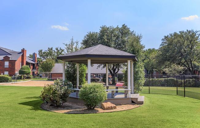 A scenic view of a gazebo surrounded by lush green grass and shrubs, with sunny skies above. The gazebo features a gray roof and white wooden structure, set in a residential area with trees in the background. A fenced area is visible, adding to the tranquil atmosphere of the outdoor space.