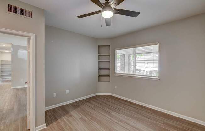 A room with a ceiling fan and light fixture, hardwood flooring, and a window with blinds.