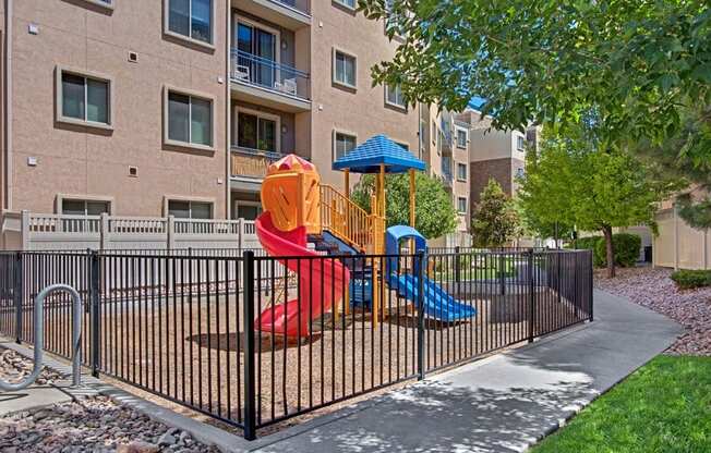 A playground courtyard here at Bridges with a colorful red and blue slide set, black safety fence, green trees, and surrounding apartment balconies, creating a bright, family-friendly outdoor space.