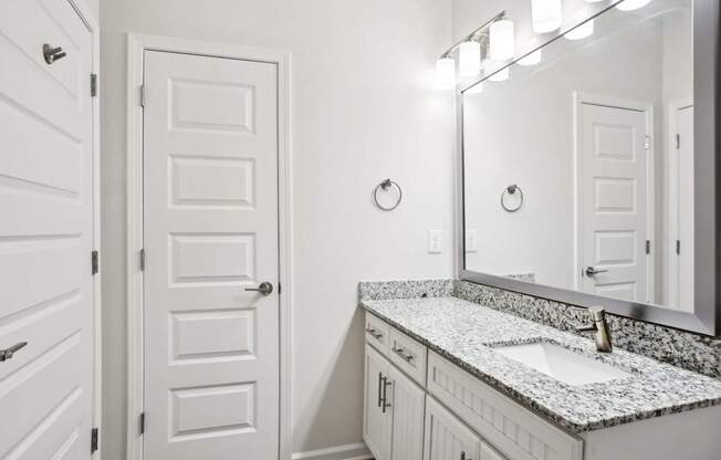 A bathroom with a white door, a marble countertop, and a mirror with lights.
