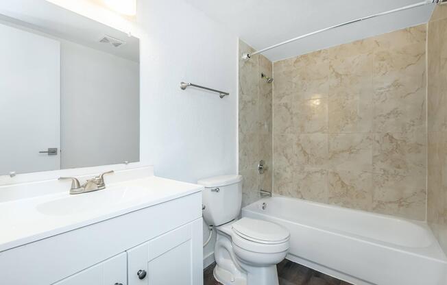 A clean and modern bathroom featuring a white vanity with a sink, a mirror above it, a toilet, and a bathtub with a shower. The walls are finished in light-colored tiles, and the flooring appears to be a dark wood finish, creating a contrast with the light walls. Natural light may be present through the door.