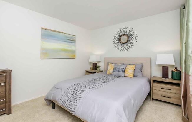 A cozy bedroom featuring a bed with a grey comforter and decorative pillows, two bedside lamps on wooden nightstands, and a round mirror on the wall. There's a colorful abstract painting above the bed and green curtains framing a window, creating a warm and inviting atmosphere.
