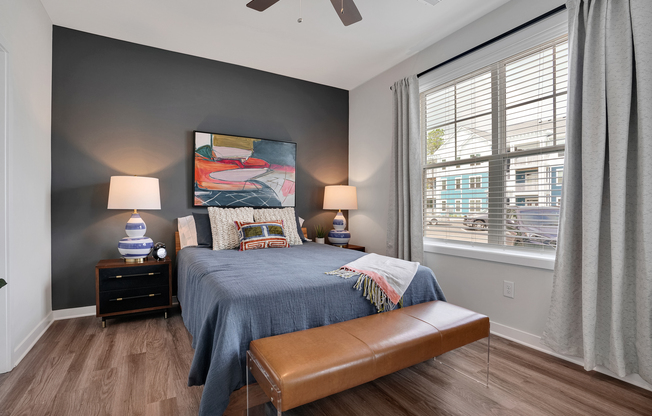 A cozy bedroom featuring a dark accent wall, a blue bedspread, and decorative pillows. There are two table lamps on either side of the bed, a stylish painting above the bed, and a decorative bench at the foot. The room has large windows with white blinds, allowing natural light to brighten the space.