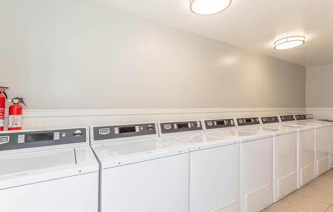 Row of white washing machines with digital control panels in a well-lit laundry room. The walls are painted a light gray, and there are two round ceiling lights illuminating the space. A fire extinguisher is mounted on the left side of the image. The floor is tiled in a neutral color.