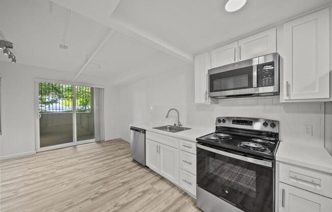 A modern kitchen featuring white cabinets, stainless steel appliances including a microwave, stove, and dishwasher, with a spacious countertop. A large window allows natural light to fill the room, complementing the light wood flooring and creating a bright, inviting atmosphere.
