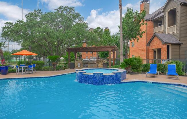 A bright outdoor pool area featuring a clear blue pool with a spa section, surrounded by lush greenery. There is a barbecue grill area under a pergola, along with several blue lounge chairs and an umbrella. In the background, there are apartment buildings and palm trees under a partly cloudy sky.