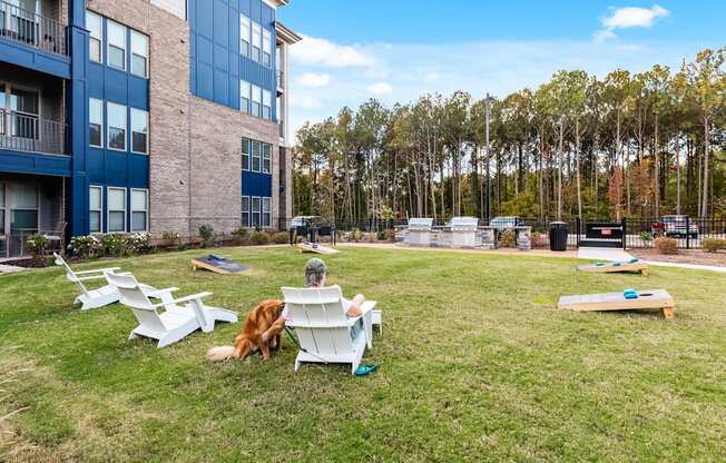 A person is sitting on a white chair in a grassy area with a dog and a picnic table.