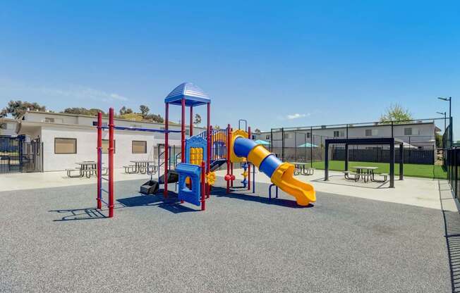 A playground with a blue and yellow slide, red and blue climbing frame, and a grey ground.