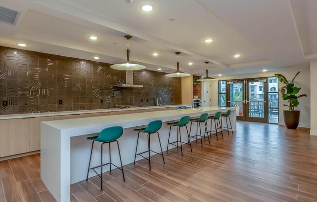 Modern kitchen with a long white countertop and high green chairs. The backsplash features stylish textured metallic tiles. Large windows provide natural light, and there is a door leading to an outdoor area. A potted plant adds a touch of greenery to the space, creating a welcoming atmosphere.