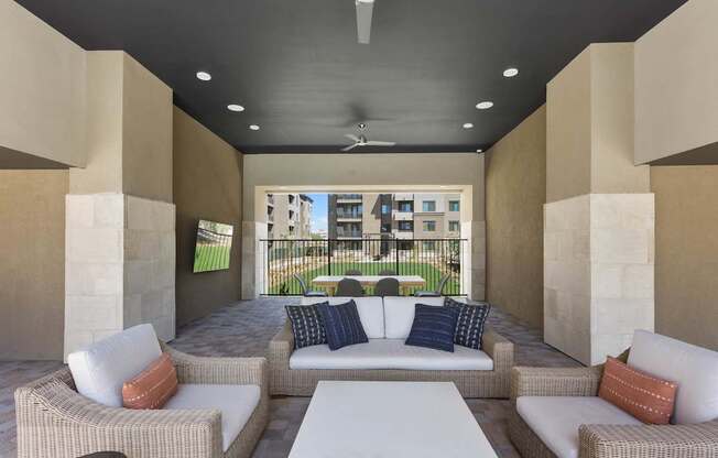 A covered outdoor lounge here at Reflect at Dobson Ranch featuring woven patio seating with plush cushions arranged around a modern coffee table, a ceiling fan and recessed lighting overhead, stone-accented columns, and an open view toward landscaped courtyard greens and surrounding apartment buildings, creating a comfortable space for relaxing or gathering outdoors.