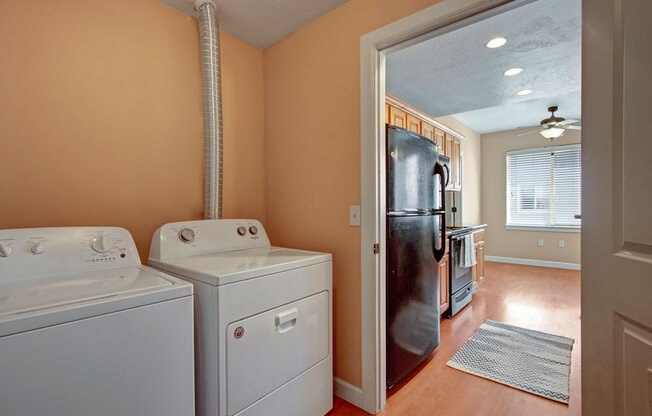 A laundry area here at Bridges with a white washer and dryer, peach-toned walls, and a vent duct, opening into a bright kitchen with wood cabinets, black refrigerator, and warm wood-look flooring.