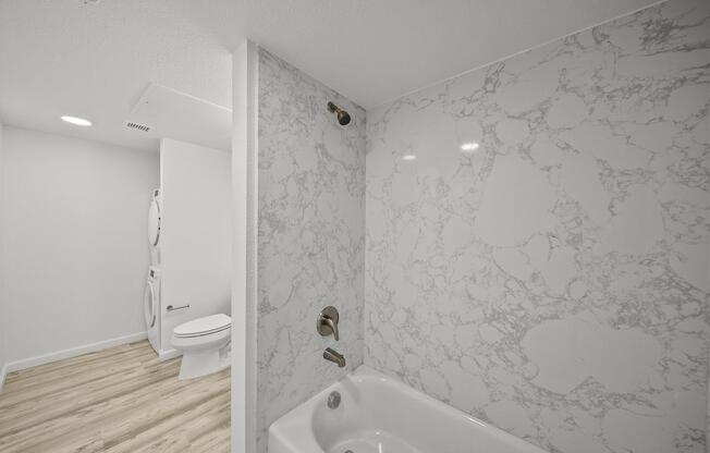 A modern bathroom featuring a bathtub with a marble-patterned wall, a gold shower fixture, and wooden flooring. In the background, a toilet is visible alongside a stackable washer and dryer unit, all in a bright, well-lit space.