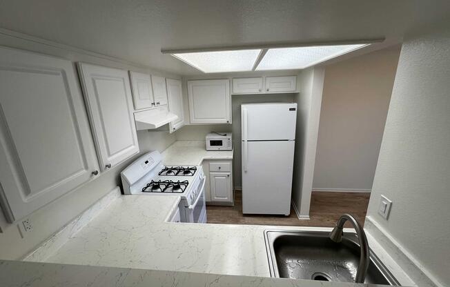 A modern kitchen view from above, featuring white cabinets, a gas stove, a microwave, and a large refrigerator. The countertops are light-colored, and a stainless steel sink is visible in the foreground. The space is well-lit with overhead lights, and the flooring appears to be wood.
