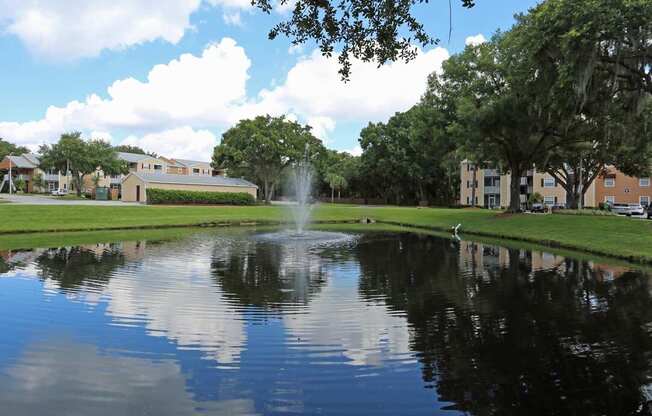 A fountain in the middle of a pond surrounded by trees and buildings.