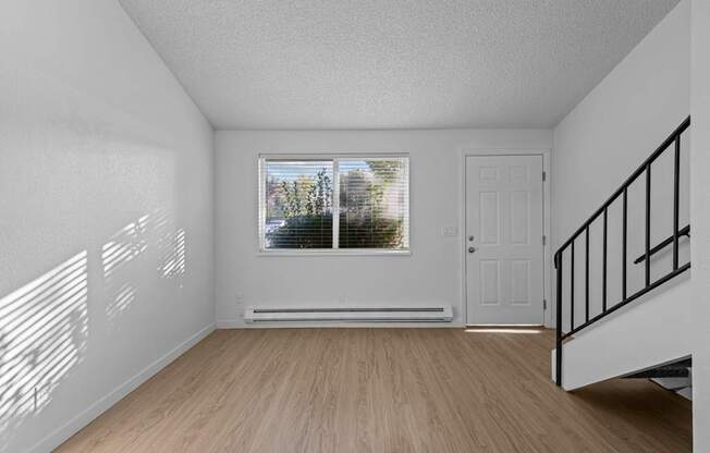 A room with a wooden floor, a window with blinds, a white door, and a staircase with a black railing.