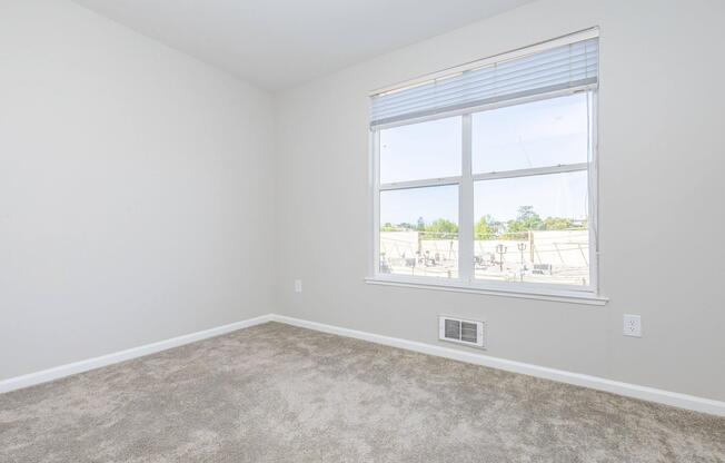An empty, well-lit room with light gray walls and a soft carpet. A large window offers a view of the outside, allowing natural light to fill the space. The room is uncluttered and features a vent near the floor.