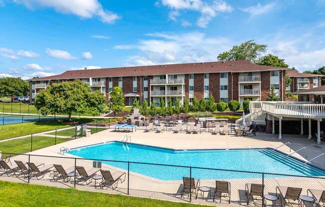 A large swimming pool surrounded by lawn chairs and umbrellas in front of a multi-story apartment building.