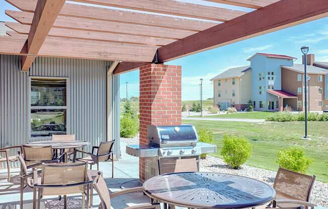 A shaded outdoor patio here at Dakota Ridge featuring a wooden pergola with exposed beams, round metal tables with mesh chairs, a stainless steel grill beside a brick column, and open views of manicured green lawns and nearby apartment buildings under a bright blue sky.