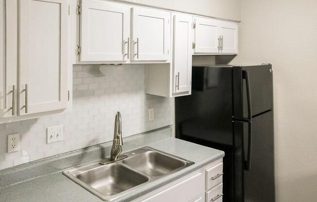 A modern kitchen featuring white cabinets, a stainless steel double sink, and a granite countertop. A black refrigerator is positioned against a light-colored wall with a white tiled backsplash. The overall design is clean and minimalist, emphasizing functionality and simplicity.