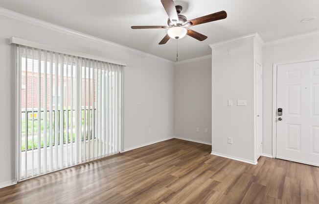 Bright and spacious room with light-colored walls and wooden flooring. A ceiling fan is mounted above, and large vertical blinds cover a window that looks out onto greenery. The entrance door is visible on the right, contributing to a welcoming atmosphere.