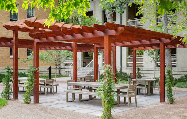 A wooden pergola with benches and tables is surrounded by greenery.