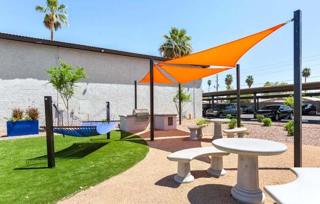A white table and chairs are set up under an orange shade sail in a sunny outdoor area.