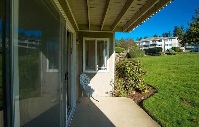 A private covered patio here at Andorra featuring a concrete outdoor space with a small decorative chair, sliding glass door entry, and an adjacent window, overlooking well-maintained green lawns, landscaped shrubs, and nearby apartment buildings, creating a peaceful outdoor extension ideal for relaxing and enjoying fresh air.