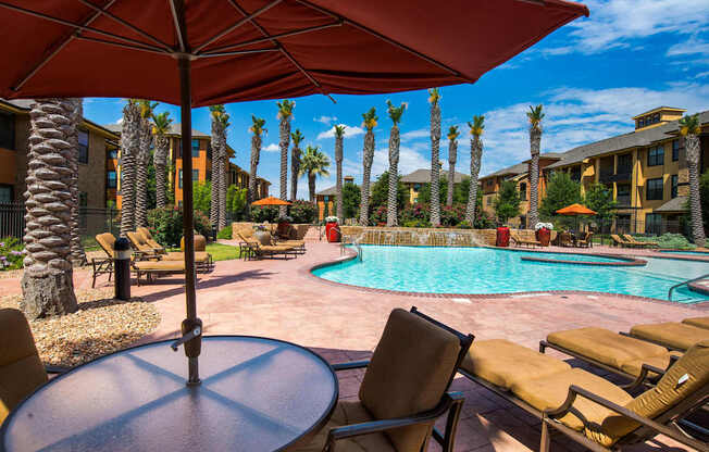 A resort-style outdoor pool area here at Tuscany at Faudree featuring a sparkling curved swimming pool surrounded by palm trees, tan lounge chairs, and shaded seating under large umbrellas. Warm-toned buildings, landscaped greenery, and a sunny blue sky create a relaxing, vacation-inspired community atmosphere.