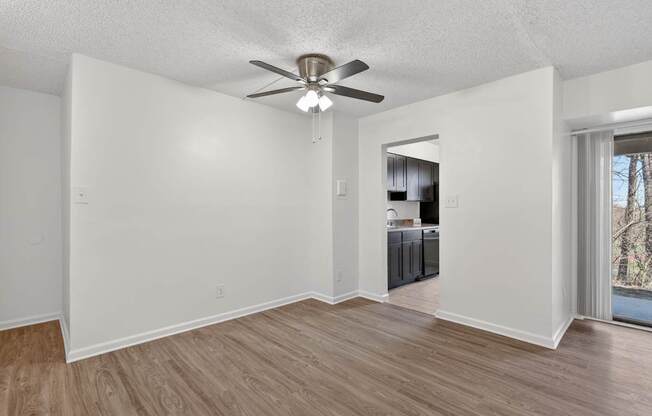 A room with a ceiling fan and light fixture, hardwood floors, and a kitchenette in the background.