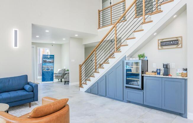 Modern interior of a spacious entryway featuring a wooden staircase with metal railings. On one side, a blue couch and a leather chair are arranged around a table. A refreshment area with a mini fridge and snacks is visible, along with a sign that reads âCoffee & Bar.â Bright, natural light illuminates the space.