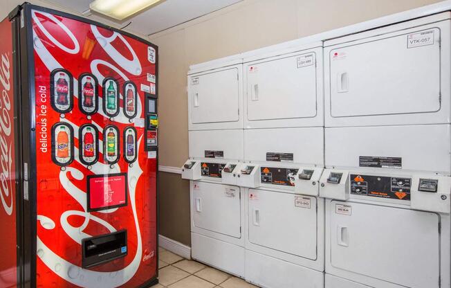 A Coca-Cola vending machine next to several white commercial washing machines and dryers in a laundry room setting. The walls are light-colored, and the flooring is tiled.
