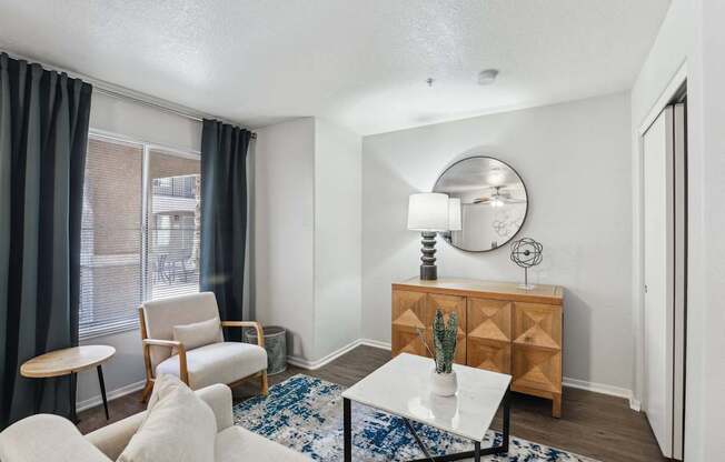 A living room with a white sofa, a wooden chair, a white coffee table, a wooden cabinet, a round mirror, and a blue and white rug.